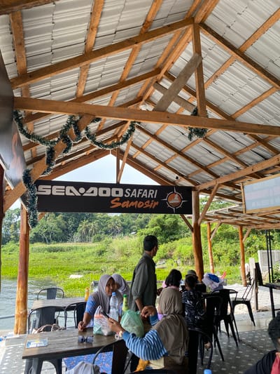 The image shows an outdoor seating area with a tin roof supported by wooden beams. In the foreground, several people are seated at wooden tables, some of whom are interacting. There is a sign visible that reads "SEADOO SAFARI SAMOSIR". In the background, there is a body of water with abundant green vegetation, suggesting a natural environment. The scene appears to be a restaurant or a recreational spot located in Tuk Tuk, Samosir Island, Indonesia, known for its natural beauty and watersports.