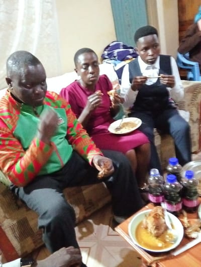 Three individuals are seated on a couch, enjoying a meal together. The person on the left wears a green and red patterned jacket, while the woman in the center is dressed in a maroon dress. The person on the right sports a white shirt and black vest. In front of them is a table with bottled drinks and a plate of food, presumably chicken in gravy. They are all actively eating, creating a scene of shared dining and socializing.