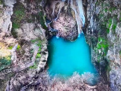The image is an aerial view of a body of water, likely a pool or small lake, with a vibrant turquoise color. Waterfalls cascade into the pool from above, and the surrounding landscape is characterized by rocky cliffs with patches of green vegetation, possibly moss or grass. Leafless trees or shrubs are clustered around the edges of the pool and along the cliffs, suggesting it may be wintertime. In the bottom left, a set of stone steps is visible, adding an element of human influence to the scene.
While the image is reminiscent of several locations with waterfalls and rock formations, there are no recognizable features to confirm the location is in Basingstoke and Deane, United Kingdom.