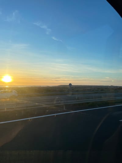 The image presents an outdoor scene at sunset, observed from a vehicle on a highway near La Palme, France. The upper two-thirds of the frame are dominated by a clear sky, transitioning from bright blue overhead to a prominent orange and yellow gradient at the horizon. The sun, a brilliant yellow orb, is positioned intensely on the far left at the horizon, casting a strong glare and reflecting brightly on the water below. A faint airplane contrail is visible in the middle-left sky.
Below the horizon, a calm body of water, likely a coastal lagoon or étang, stretches across the mid-ground, mirroring the sky's colors and the sun's brilliance. Beyond this water, a low-lying, vegetated landscape extends horizontally, characterized by scrubland and distant, slightly elevated terrain on the right. A circular road sign, featuring a white face and a dark border, stands within this vegetated area on the right side of the highway.
In the foreground, a multi-lane asphalt highway runs from left to right, parallel to the horizon. The dark asphalt surface displays white lane markings, including a solid white line defining the shoulder. A metal crash barrier, consisting of two horizontal rails, separates the highway from the adjacent landscape. A light-colored (possibly white or silver) SUV or crossover vehicle is partially visible on the far left of the highway, traveling in the same direction as the vantage point, illuminated by the setting sun. Reflections, indicative of a window, are visible across the lower portion of the image, including a dark, amorphous shape in the lower right corner. No individuals are explicitly discernible.