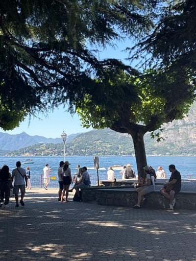In Gittana, Italy, a paved walkway lined with trees overlooks a wide, calm lake. Several individuals are present. On the left, a couple walks away from the camera. Further along the walkway, a group of people stand near a decorative lamp post, facing the lake. Some are engaged in conversation, while one individual appears to be taking a photograph. In the foreground, under the shade of a large tree, several people are seated on a stone wall. A woman in the foreground on the right is seated on the wall, wearing a white top and denim shorts, holding a bag of chips. A man is seated next to her, wearing a dark polo shirt and shorts. The lake is dotted with a few boats, and the distant shore is lined with buildings and lush green hills that rise into mountains under a clear blue sky. The scene appears to be a public area, possibly a promenade or park, enjoyed by visitors on a sunny day.