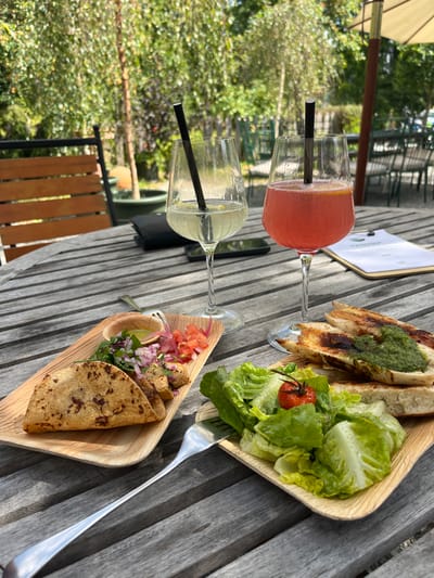 The image depicts an outdoor dining scene on a slatted wooden table, likely at a restaurant or cafe patio in Kitzbühel, Austria. In the foreground, two stemmed glasses are present: one on the left contains a light yellow beverage with a black straw and citrus garnish, and one on the right holds a reddish-orange beverage with a black straw and orange slice.
To the left, a rectangular light-brown plate holds two folded, browned flatbreads or tacos. Adjacent to these are distinct piles of diced red onion, chopped red tomato, green herbs, and a small cup of light green sauce. A second rectangular light-brown plate on the right features several slices of toasted bread topped with a green pesto-like spread, accompanied by a green salad comprising lettuce leaves and a single red cherry tomato. A silver dining fork rests on the table, partially under the salad plate.
Between the two beverages, a dark-colored wallet or clutch and a black smartphone are visible on the table. A white clipboard or menu with green text is partially visible on the far right. The background consists of blurred green foliage, indicative of trees or shrubs, and indistinct outlines of additional outdoor seating, suggesting a garden or patio setting during daylight hours. No individuals are present in the scene.
