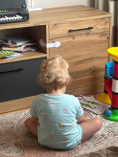 A young child with blonde, curly hair, viewed from behind, sits on a rug in front of a wooden cabinet. The child wears a light blue t-shirt with pink and yellow flower patterns. The cabinet has a light brown wood grain finish with a black handle on one drawer. A white safety latch is attached to the cabinet door. On the top shelf of the cabinet, to the left, is a black toy keyboard. To the right of the cabinet, a colorful toy structure with red, yellow, and blue components is visible. The floor is covered with a textured rug. A book with illustrations of green crocodiles is on the rug in front of the child. The scene appears to be indoors, possibly in a home environment. There is no discernible information about the specific location within Krāslava, Latvia.