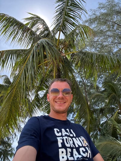 In Ban Khuk Khak, Thailand, a man with short brown hair and a beard is captured in a selfie. He is wearing sunglasses with reflective lenses and a dark blue t-shirt with white lettering that reads "CALI FORNIA BEACH". The man is smiling and positioned in front of lush green palm trees, with sunlight filtering through the fronds. The sky is visible in the background, appearing bright and clear. The setting suggests a tropical or coastal environment.