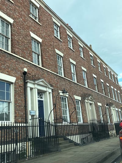 The image shows the exterior of a tall brick building with many windows. The perspective is from a low angle, looking up towards the sky. The building's facade is made of red bricks, and there are white accents around the windows. The windows appear to be white-framed and reflective. Some of the windows have a diamond pattern in their glass. A black metal fence runs along the lower part of the building, adjacent to a stone pathway. The sky above is overcast and a pale blue.
There are no people visible in the image. Several black metal lamp fixtures are attached to the building at different levels, some with glass globes that appear to be unlit. One notable detail is a sign with the text "bartletts" visible on the building near the fence.
The scene appears to be outdoors, in an urban or suburban setting, given the architecture of the building and the presence of a pathway and fence. The lighting suggests it is daytime, possibly overcast, as there are no strong shadows. The overall mood is neutral and observational. The tilted angle of the photograph adds a dynamic feel to the otherwise static architectural subject.