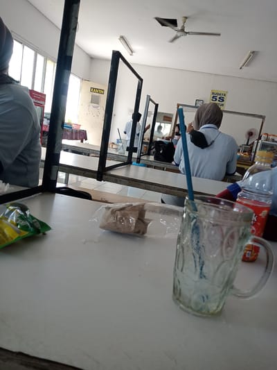 The image depicts a scene in what appears to be a canteen or cafeteria, likely located in Nglundo, Indonesia. There are several people, including women wearing hijabs, sitting at tables. The tables are separated by black metal frames. There's a glass with a straw on the table in the foreground, along with an open snack bag and a plastic bag. In the background, a door labeled "KANTIN" is visible. A poster about "BUDAYA 5S SENYUM" is on the wall. A ceiling fan is also visible.