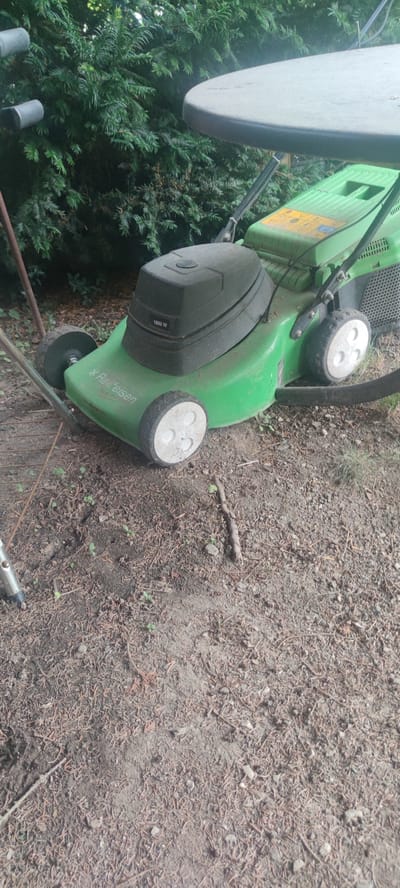 This is a medium shot, eye-level photograph of a green electric lawnmower parked on a dirt and pine needle-covered ground. The lawnmower is the central subject, with its black engine cover prominently displayed. Visible text on the engine cover reads "1800 W". The side of the lawnmower has the brand name "Raiffeisen" and the model name "AL-KO" written on it. The lawnmower has four wheels, two larger ones at the back and two smaller ones at the front. A portion of a large, light-colored, round object, possibly a table, is visible in the upper right corner of the frame, casting a shadow. Behind the lawnmower, there is a dense green hedge or evergreen shrubbery. The ground is a mix of soil, dry pine needles, small twigs, and pebbles, with a few small green sprouts of grass or weeds visible. A metal pole, part of some outdoor equipment, is partially visible on the left side of the frame. The lighting suggests it is daytime, with natural light illuminating the scene. The overall setting appears to be an outdoor garden or yard, likely in a residential area in Liebenburg, Germany, given the context. No people are present in the image. The scene is static, with the lawnmower appearing to be unused at the moment. The image conveys a sense of everyday outdoor equipment in a natural setting.