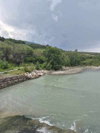 The image shows a view of the Karatal River flowing through the city of Tekeli, Kazakhstan. The river is a pale green color and appears to be moderately deep. On the left bank of the river, a gabion wall is constructed, reinforcing the riverbank with stones contained in wire cages. Beyond the gabion wall, a paved pathway runs parallel to the river, bordered by lush green vegetation and trees. The riverbank on the right is composed of a rocky shore, with a mix of small and medium-sized stones. Further inland, rolling hills covered in dense green trees and vegetation rise towards a cloudy sky. The sky is overcast with heavy grey clouds, suggesting a possibility of rain. Power lines are visible stretched across the upper part of the frame, suggesting proximity to infrastructure. There are no people or animals visible in the image. The overall scene depicts a natural landscape integrated with man-made structures for riverbank protection, under a moody sky.