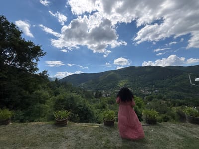 A woman with long, dark, curly hair stands with her back to the camera, wearing a long, flowing, pink dress with a subtle pattern. She is looking out over a vast, verdant valley filled with rolling hills covered in lush green trees. In the distance, a small village is nestled amongst the trees, with buildings visible amongst the foliage. The sky above is a vibrant blue, dotted with dramatic, fluffy white clouds that cast shadows on the landscape.
The setting appears to be an elevated viewpoint, possibly a terrace or balcony, with a grassy foreground. Two large potted plants, filled with spiky green foliage, flank the woman. A yellow hose or cable lies on the grass between the pots. The overall atmosphere is peaceful and serene, with a sense of awe inspired by the expansive natural scenery. The time of day appears to be daytime, with bright sunlight illuminating the scene and creating a contrast between light and shadow. The weather seems to be fair, with a mix of sun and clouds. The location is identified as Lukovo, Bulgaria. No text is visible in the image.