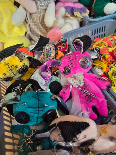 The image depicts a collection of handmade plush toys, commonly referred to as "creepy dolls," arranged within a light gray plastic basket. The dolls are characterized by their somewhat unsettling aesthetic, featuring mismatched button eyes, stitched facial features, and exposed seams. One prominent doll is bright pink, adorned with large, reflective button eyes, stitch marks, and a delicate butterfly charm hanging from a chain around its neck. Another doll, teal in color, also has button eyes and a star-shaped charm dangling from its neck. Other dolls visible in the image are yellow and feature similar construction. The dolls are interspersed with various fabric scraps and embellishments, including safety pins, ribbons, and translucent mesh. The background suggests a cluttered environment, with other plush toys and fabric items partially visible. The lighting is moderate, casting soft shadows on the subjects. The overall scene appears to be an indoor setting, possibly a craft area or a display of handmade items. There is no discernible information in the image that can indicate its location in the city of Красный Ключ, Russia.