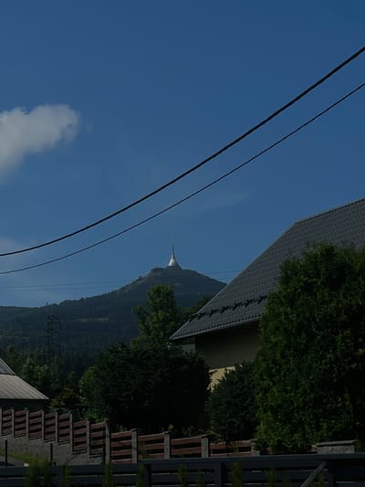 The image captures a view of the Ještěd Tower, a distinctive telecommunications tower and hotel situated atop Ještěd Mountain, overlooking the city of Liberec, Czechia. In the foreground, a portion of a building's grey, corrugated roof is visible, along with dense green coniferous trees. Further back, a wooden fence with stone pillars runs horizontally across the frame, separating properties. Beyond the fence, a densely forested mountainside rises, dotted with trees. A power line runs diagonally across the clear blue sky, with a single white cloud to the left. The Ještěd Tower, with its unique tapered design, stands prominently on the peak of the mountain in the distance, its white structure contrasting with the green and grey hues of the landscape. The overall scene depicts a sunny day in a scenic, semi-rural setting with the iconic landmark of Liberec visible.