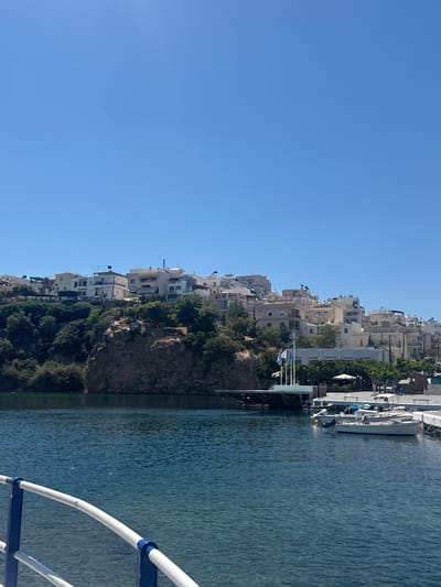 This image depicts a view of a coastal town built on a rocky hillside overlooking the sea. The town features numerous multi-story buildings, mostly white or light-colored, with balconies and varying architectural styles. Green trees and vegetation are interspersed among the buildings, clinging to the steep terrain.

In the foreground, a portion of a white railing with blue accents suggests the vantage point is from a boat or a waterfront structure. The sea is a deep blue-green, with gentle ripples on its surface. A white boat is docked at a pier or quay on the right side of the image.

A few people are visible near the water's edge by the boat, one appearing to be wearing a yellow hard hat. Another individual is leaning over the side of the boat. The overall scene suggests a sunny day, likely in the morning or afternoon, given the clear blue sky and bright lighting. The location is identified as Agios Nikolaos Municipal Unit, Greece, which aligns with the Mediterranean coastal architecture and landscape. There is no visible text in the image.
