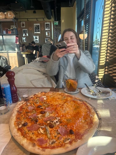 The image displays an indoor scene within a restaurant in Mysiadło, Poland. In the foreground, a large, partially consumed pizza dominates a light-colored, patterned table. The pizza features distinct toppings including ham slices, mushrooms, olives, and artichoke pieces, applied over a red sauce and cheese base. To the right of the pizza, a transparent glass mug containing a hot beverage with fruit slices (likely lemon or orange) rests on a white saucer. Adjacent to the mug is a white plate holding a fork, a knife, and a piece of bread crust, indicating prior consumption. A red pepper grinder and a blue salt shaker are also positioned on the table, along with two glass votive candle holders, one containing a lit candle.
Seated behind the table, a female individual, approximately 25-35 years old, is shown smiling towards the viewer. She wears a light gray cardigan over a white top and holds a smartphone horizontally with both hands, seemingly capturing an image or video of the pizza. A silver ring is visible on her left hand. A beige, textured coat is draped over a chair or booth seat to her left.
The background reveals a bustling restaurant interior. Several blurred figures are visible, seated at other tables. The back wall is yellow and decorated with multiple framed pictures. A wooden shelving unit on the left holds various bottles and jars, along with decorative items marked with an 'L'. A green illuminated exit sign is mounted high on the back wall. Large windows on the right side of the frame allow bright sunlight to enter, casting strong, defined shadows from horizontal blinds across the interior surfaces. The exterior view through the windows suggests a parking area or street under a clear blue sky.