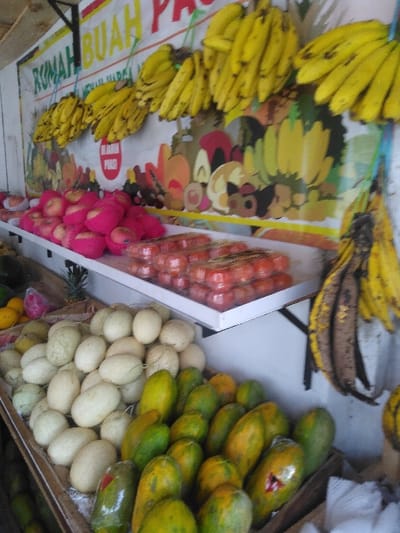 A vibrant fruit stall, located in the city of Yae Tha Yauk, Myanmar, is presented in an eye-level shot. The background features a large white banner adorned with illustrations of various fruits and text, including 'RUMAH BUAH PA...' and 'DI JAMIN PUAS!' on a red rectangle. Numerous bunches of ripe yellow bananas, some with brown spots, hang from ropes against this banner, with several older, darkened bananas also hanging on the far right. Below the banner, a white two-tiered shelving unit displays additional produce. The upper shelf on the left holds red apples, many individually encased in pink protective foam netting. To the right, clear plastic clamshell containers are stacked, each holding small, round orange-red fruits, possibly tomatoes or plums. The lower level, constructed from wooden crates or bins, features a large pile of light green to cream-colored melons on the left. Adjacent to these, on the right, is a substantial display of green and yellow-orange papayas, one with a red triangular sticker. A pineapple is partially visible behind the melons on the far left. No people are visible.