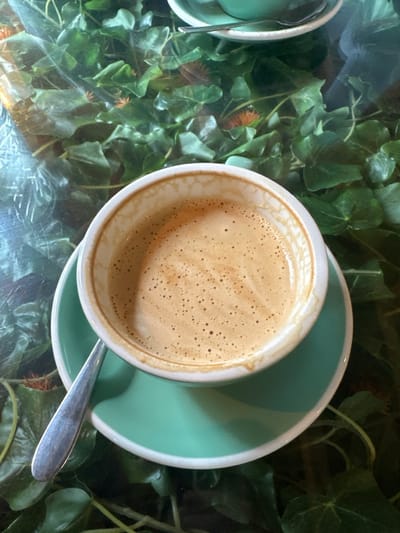 An overhead view captures a white ceramic coffee cup containing a light brown beverage with visible crema and microfoam, centered on a light turquoise/mint green saucer. A silver spoon is positioned beneath the cup, resting on the saucer. This assembly sits on a clear glass table. Beneath the glass, a decorative arrangement of artificial green ivy leaves is visible, interspersed with small, spiky, orange-brown floral or botanical elements. A portion of a second identical cup and saucer set is discernible in the upper right background. No individuals are depicted. The specific location within Salisbury, United Kingdom, cannot be determined from the visual information provided.