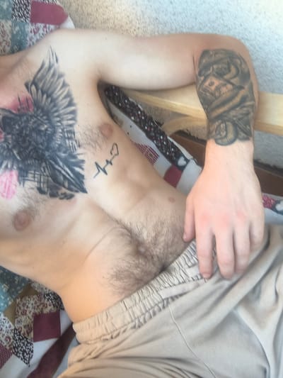 The image shows a close-up, overhead view of a person lying down. The person has a tattoo of an eagle or raven on their chest and a small tattoo of a heartbeat line with a heart on their abdomen. They are wearing light-colored sweatpants with an elastic waistband. The person's right arm, which is visible on the right side of the frame, also has a tattoo depicting a stack of money and possibly a hand. The background suggests a bedroom or living area, with a colorful, patterned blanket or quilt visible behind the person's upper body and a textured floor. The lighting is soft, indicating it could be daytime or a well-lit indoor space. There is no visible text in the image. The setting appears to be casual and relaxed.