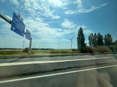 A view from a vehicle on a highway in Graincourt-lès-Havrincourt, France, on a clear, sunny day. The sky is a vibrant blue, dotted with fluffy white clouds. The sun is high, indicating midday. The landscape features a grassy field with several wind turbines spread across it, their blades silhouetted against the bright sky. Some trees are visible on the right side of the frame.

In the foreground, a concrete barrier separates the highway lanes. The road surface is asphalt, marked with white lines. A metal guardrail runs alongside the road.

There are two blue road signs visible on the left, mounted on a pole. The upper sign, partially obscured, has "A2" and "E19" printed on it. The larger, lower sign displays directional information: "LIEGE", "BRUXELLES", "VALENCIENNES", and "CAMBRAI", with white arrows pointing downwards, indicating the direction of travel. Below this, a smaller sign indicates "A26 E17" with "REIMS" and "ST QUENTIN" and a downward arrow.

The scene suggests travel on a major road, likely a French autoroute, given the signage. The presence of wind turbines indicates a rural or semi-rural area, possibly an area known for renewable energy production. No people are visible in the image. The overall mood is calm and peaceful, characteristic of a pleasant day for driving.