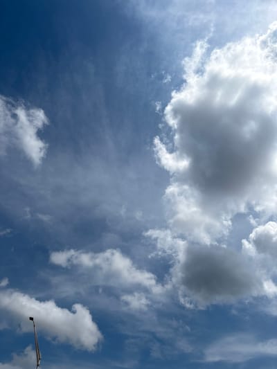 A wide-angle, vertically oriented, medium shot captures a bright, cloudy blue sky from a low-angle perspective. Dominating the right side of the frame, a large, dense cumulus cloud formation features stark white highlights and deep gray shadows, indicating significant depth. Smaller, wispy clouds, appearing as white streaks, are scattered across the upper left and lower sections of the image. The lower left corner shows the top of a metal lamppost, its angled shaft extending upwards towards the sky. The scene is likely outdoors in Duette, United States, given the clear, expansive sky and the presence of a lamppost, consistent with an outdoor setting.