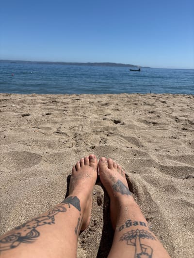 The image presents a low-angle, first-person perspective captured from a sandy beach in Tomé, Chile. In the immediate foreground, the bare feet and lower legs of an individual are visible, partially covered with light brown sand. Both lower legs are extensively tattooed with black ink designs, featuring various patterns, abstract shapes, and visible text fragments. The middle ground consists of a calm, blue body of water. A small orange buoy is present in the water on the left side of the frame. Towards the center-right, a small, dark-colored boat, likely motorized, is observed on the water, with a single occupant appearing to operate it. A low, dark landmass stretches across the distant horizon. The upper portion of the image is dominated by a clear, bright blue sky, indicating a sunny day. Shadows from the person's limbs are cast onto the sand.