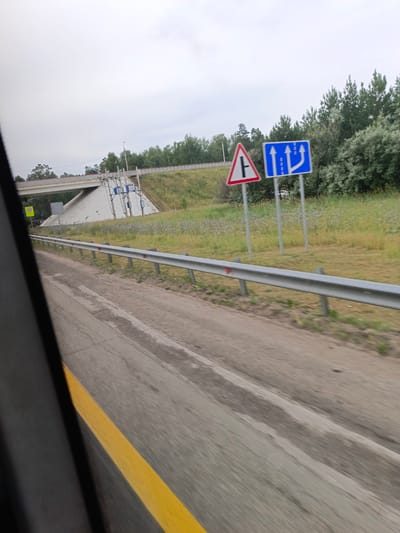 A road scene in Sibirsky, Russia, features a highway overpass with a concrete barrier and a grassy slope. Several road signs are visible: a triangular sign with a black "T" shape on a white background with a red border, indicating a junction or intersection, and a blue rectangular sign with white arrows indicating lane guidance for traffic merging or changing direction. A yellow traffic line runs along the road. Trees and vegetation line the embankment of the overpass. The sky is overcast. The image appears to be taken from a moving vehicle.