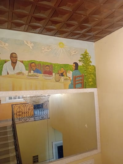The image depicts a mural on a wall, likely in a residential or public building in Douala V, Cameroon. The mural, painted in a somewhat naive style, portrays a family gathering around a table, with figures resembling adults and children. Above them, stylized cherubic figures and a sun with rays are painted, suggesting a celestial or heavenly theme. The mural is positioned above a large, ornate mirror that reflects a staircase with a decorative metal railing and a chandelier. The overall environment appears to be an interior space with yellow-painted walls and a textured, dark brown ceiling. The scene captured by the mural suggests a peaceful and perhaps celebratory moment, possibly a family meal or religious gathering.