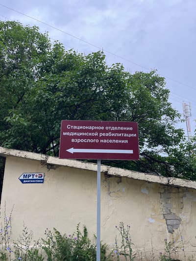 The image shows a street sign in the city of Samara, Russia. The sign, which is a deep red color with white text, reads "Стационарное отделение медицинской реабилитации взрослого населения" (Inpatient Department of Medical Rehabilitation for the Adult Population). An arrow points to the left, indicating the direction to this department. Below this sign, on a beige wall, is another smaller sign with a blue arrow pointing left that reads "MPT+ diagnostic" in Russian. This suggests the presence of an MRI diagnostic center. The scene is outdoors, with lush green trees in the background and an overcast sky. A weathered beige wall with some exposed brick is visible in the lower half of the image. In the far background, a telecommunication tower can be seen. Wildflowers are growing at the base of the wall.