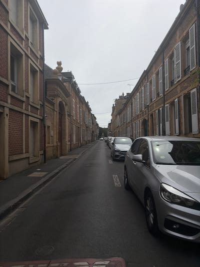 This street scene in Charleville-Mézières, France, shows a narrow road flanked by buildings of brick and stone construction. The buildings are several stories high with various architectural details, including ornate window frames, dormers, and what appear to be closed shutters. Parked cars line one side of the street, primarily silver or grey in color. The road surface is asphalt, wet, and marked with parking designations. The sky is overcast, and the overall impression is of an urban environment under grey weather conditions. The buildings display a mixture of red brick and beige stonework, with visible piping and decorative elements.