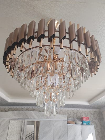 The image shows a close-up, low-angle view of a large, multi-tiered chandelier hanging from a textured white ceiling. The chandelier features a circular frame with numerous hanging crystal pendants. The upper tier of the chandelier has a series of bronze-colored, curved metal shades alternating with gold-colored cylindrical accents. The lower tier is comprised of multiple strands of clear, faceted crystal prisms. The light fixture appears to be made of metal with a gold and bronze finish. Below the chandelier, a sliver of a room is visible, including wallpaper with a floral pattern, white cabinets with a marble-like finish, and some stacked boxes on a shelf. There are no people or animals present. The location is not discernible from the image itself; the provided location of Социализм, Kyrgyzstan is assumed context and not visually verifiable.