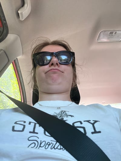 A medium shot captures a young person wearing sunglasses and a white t-shirt with the word "STUSSY" printed across the chest, seated in the passenger seat of a car. The person's head is tilted back, looking upwards towards the car's interior. A black seatbelt crosses diagonally over their chest. The background shows the car's ceiling and a window with green foliage visible outside. The image is cropped at the top and bottom, with the person's chin and chest prominent in the frame. No specific location in Cannon Beach is identifiable from the image.