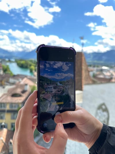 A person is holding a smartphone in landscape orientation, framing a scenic view of Thun, Switzerland. The phone displays a photo capture interface, with a grid overlay on the image being viewed, suggesting the user is composing a photograph. The captured image shows a picturesque scene of red-tiled rooftops and a river, with lush green hills in the background under a bright blue sky dotted with fluffy white clouds.

The phone itself has a purple case, and the person's hands, with manicured fingernails, are visible as they operate the device. The individual's clothing, a dark jacket, is visible at the top right of the frame. The background suggests an elevated vantage point, likely from a balcony or window, offering a panoramic vista of the town and its natural surroundings. The lighting indicates it is daytime, likely mid-morning or afternoon, with sunny weather.

Text visible on the phone screen includes "СТУДІЙНЕ СВІТЛО" (Studio Light) and "ФОТО" (Photo), indicating the camera mode and some available settings. The overall mood is one of relaxed enjoyment and appreciation of the beautiful scenery.