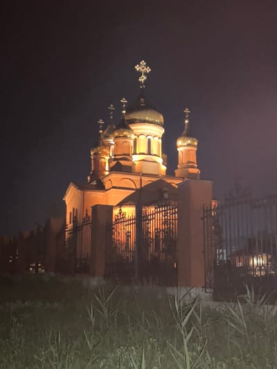 The image shows an illuminated orthodox church at night, likely in the city of Novoteroksky, Russia. The church features several golden domes topped with crosses, and the building itself is a warm, orange hue from the lighting. A decorative black metal fence surrounds the church, with some vegetation visible in the foreground and beyond the fence. The sky is dark, indicating nighttime, and the overall scene is peaceful and serene. There are no people visible in the image.
