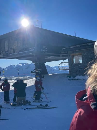 The image captures a vibrant, sunny day at a ski resort, rotated 90 degrees clockwise from its original orientation. The perspective is first-person, looking down past a red jacket with a fur-lined hood.
The primary subject is a modern ski lift station, dark grey or black, with a stone-clad building section. Visible text on the station clearly reads "La Rosière" accompanied by a sun logo, "Espace San Bernardo," and "MONT VALAIS." The sun is prominently featured on the left, emitting a strong glare and lens flare against a clear, bright blue sky.
In the foreground and mid-ground, several individuals are visible in full ski and snowboard attire. Many wear red jackets, while one person is in a dark jacket and tan pants, equipped with a snowboard. Others are on skis, holding ski poles. They appear to be queuing for or standing near the ski lift, ready to ascend the mountain.
The setting is a vast snow-covered mountain landscape, with distant peaks visible under the brilliant blue sky. The bright conditions suggest it is midday, perfect for winter sports. The scene conveys an active and picturesque environment typical of a high-altitude ski destination like La Rosière in Séez, France, which is part of the Espace San Bernardo ski area.