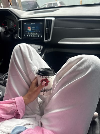 A person's hand holds a paper coffee cup inside a car. The cup has a black lid and a white body with a graphic of a city skyline and a red flower, along with the text "Ekaterinburg". The person is wearing white pants and a pink shirt. The car's dashboard and media screen are visible in the background. The location is in the city of Sovkhozny, Russia.