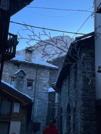 The image captures a winter scene in a mountain village, likely Courmayeur, Italy, as indicated by the context. The photograph is oriented vertically (rotated 90 degrees counter-clockwise from its natural state), showing a view upwards through a narrow street or passage.

In the corrected orientation, the scene depicts stone and wooden buildings lining the left and bottom edges of the frame, some with snow accumulated on their dark roofs. One building on the left features a stone chimney and small windows. Overhead, numerous wires and cables crisscross the sky, alongside a decorative structure resembling illuminated branches or string lights, suggesting a festive or winter ambiance.

The background reveals a magnificent snow-covered mountain peak bathed in bright sunlight, which creates a prominent lens flare in the upper-left sky (after correction). The sky itself is a clear, bright blue. A small portion of a vibrant red object, possibly a person's clothing or another decoration, is visible on the far right.

On the wooden facade of one of the buildings, a white sign is visible, displaying "CERTIFICATO DI ECCELLENZA" (Certificate of Excellence) and the "TRIPADVISOR" logo with "2017," along with a "q-cert" logo. Another small sign indicates "Wi-Fi." The image evokes a sense of peaceful, clear winter weather in an Alpine village.