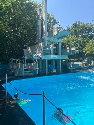 The image depicts a sunny day at an outdoor swimming pool in Rheinbach, Germany. A diving tower dominates the background, with a young male mid-flip as he jumps from one of the platforms. Several other individuals, including children, are visible around the pool. One child in green swimming trunks swims in the foreground, while another in a pink swimsuit is further in the pool. The pool water is a vibrant blue, with splashes suggesting recent activity. The surrounding area features lush green trees and wooden decking around the pool's edge.