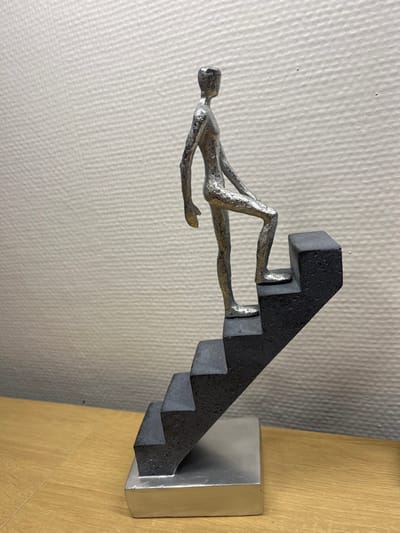 An indoor scene displays a metallic sculpture situated on a light brown wooden surface, positioned against a light gray wall with a subtle textured pattern. The sculpture features a stylized, silver-colored human figure, rough in texture and lacking distinct facial features, depicted in the act of ascending a dark gray, five-step staircase. The figure's right foot rests on the second step from the top, with its left foot on the step below, conveying a sense of upward movement. The dark gray staircase structure is secured to a rectangular silver-colored base. The wooden surface exhibits visible grain, and the background wall presents an even, fine texture. While the location is indicated as Schönau am Königssee, Germany, the image contains no discernible visual elements to confirm this specific geographic placement.
