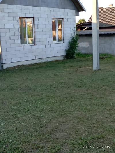A construction site, possibly in the city of Урыцкі сельскі Савет, Belarus, is captured in an outdoor, daytime photograph. The foreground is dominated by a freshly mowed lawn, with patches of dry, light brown grass interspersed with greener areas. A light grey concrete pole stands prominently in the mid-ground, slightly off-center to the right. Behind the pole, the scene transitions to a partially constructed building with white concrete blocks as its walls. Two windows are visible on this building, each with white frames and glass reflecting the surrounding environment. To the right of the building, a grey concrete fence runs horizontally across the image, and beyond that, another building with a dark, corrugated roof is visible. The sky above is clear and bright, suggesting fair weather. A date and time stamp at the bottom right corner reads "20.06.2026 20:19".