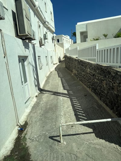 The image depicts a narrow, outdoor alleyway in Thira Municipal Unit, Greece, characterized by whitewashed buildings and a bright blue sky. On the left, a white building features several air conditioning units mounted on its exterior and a white door with a glass pane. The building's facade is textured and shows some discoloration along the base. The alley floor is paved with concrete that appears worn and cracked in places. A black stone wall runs along the right side of the alley, topped by a white slatted fence. Sunlight casts sharp, linear shadows across the alley floor, originating from the fence. In the foreground, a rusty, white metal barrier is present on the ground. Near the base of the white building, a discarded blue and white aluminum can and some cigarette butts are visible on the ground, near a patch of sparse green grass. The overall scene suggests a typical residential or commercial area in a Mediterranean town.