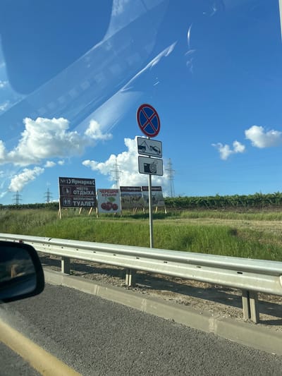 This image depicts a roadside scene on a sunny day, likely near Primorskii, Russia, based on the signs. The perspective is from inside a vehicle, looking out towards a grassy field with several large billboards.

The billboards advertise what appears to be a market or fair. One prominent sign reads "№1 Ярмарка" (No. 1 Fair) and indicates "МЕСТО ОТДЫХА" (Rest Area) and "БЕСПЛАТНЫЙ ТУАЛЕТ" (Free Toilet). Another sign features an image of cherries and the text "ЧЕРЕШНЯ КУБАНИ" (Cherries of Kuban). Further signs show images of animals like camels and a deer.

Above the billboards, a traffic sign with a red circle and a blue diagonal cross indicates no stopping or parking. Below this, two more signs show a tow truck and a camera icon, suggesting potential towing and surveillance.

The environment includes a clear blue sky with scattered white clouds. In the background, power lines and tall pylons are visible, along with rows of grapevines or similar crops. A metal guardrail runs along the edge of the road in the foreground. The road itself is asphalt with a yellow dividing line. A car's side mirror is visible in the lower left corner of the frame.