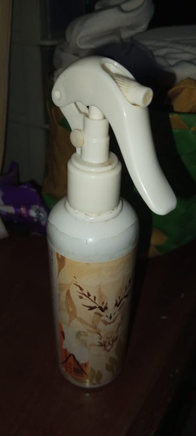 A white plastic spray bottle with a white spray nozzle sits on a dark surface. The bottle's label features a design with brown branches and leaves on a beige background. The bottle appears slightly worn, with some dirt visible around the nozzle and top rim. The background is cluttered with various objects, including fabrics and containers in muted colors. The lighting is dim, casting shadows that obscure details in the background.