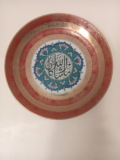 The image shows a decorative plate, featuring Arabic calligraphy in the center. The calligraphy is in black ink on a white background surrounded by an intricate blue, green, and red floral pattern. The outer rim of the plate is decorated with alternating bands of reddish-brown and gold with floral patterns. The plate is positioned on a white surface. The origin of the plate cannot be determined based solely on the image. Therefore, the location of Ksar-el-Kebir, Morocco, cannot be confirmed.
