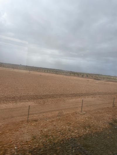 A landscape featuring a flat, barren field stretches from the center to the right, transitioning to a rocky, shrub-covered area on the extreme right. Sparse vegetation borders the field's edge, contrasting with the flat, beige expanse. Vertical posts, possibly utility or fence, are intermittently placed along the field. Above, a vast, overcast sky dominates the left portion, gradually merging into the landscape. A faint reflection hints at a window, implying the image was taken from inside a vehicle.