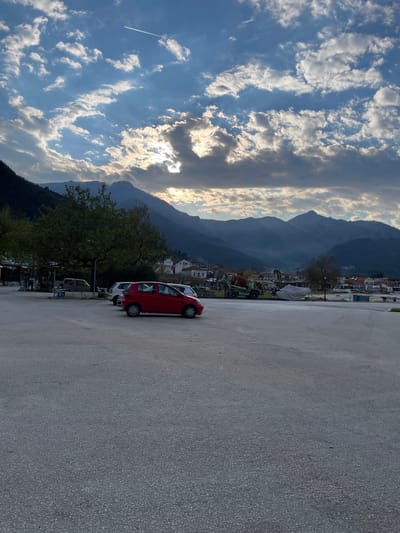 The image shows a mountain landscape in Skala Potamias, Greece. A red car is parked on a concrete area, with mountains and buildings visible in the background. The sky is blue with white clouds. A contrail from an airplane is also visible in the sky.