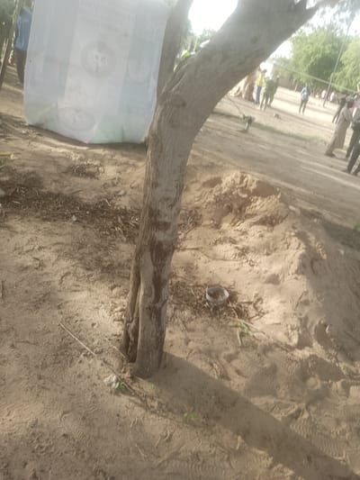 In Majemu, Nigeria, an outdoor scene unfolds with a focus on a large, white, possibly plastic sack placed on the ground near a tree. The sack displays text and circular emblems, suggesting it may contain a product or be part of a campaign. Several people are visible in the background, some appearing to be engaged in activities or conversation. The environment is sandy with sparse vegetation, including a tree with a textured trunk that dominates the foreground. The ground is uneven, with piles of sand and scattered debris. The lighting suggests it is daytime, casting shadows across the scene.