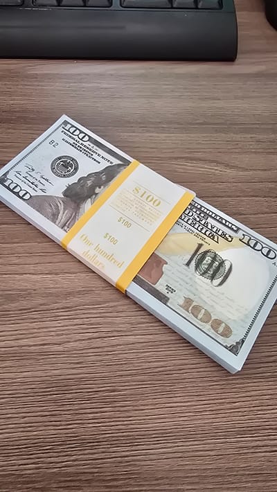 A stack of counterfeit US $100 bills is laid on a wooden desk. The bills are banded with a yellow wrapper that reads "$100" and "One hundred dollars". A black keyboard is visible in the background. The image is taken from a slightly high angle, looking down at the stack of money.