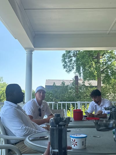 Three men are seated at a table on a covered porch. The man on the left is Black, wearing a white button-down shirt and tan pants. He is looking to his left. In the middle, a White man wearing a white polo shirt and a white baseball cap is looking forward. To the right, a White man with dark hair and glasses, wearing a white button-down shirt, is looking down at his phone. The table has a red container, a small tub of ice cream with a floral pattern, and a phone. In the background, there are trees and a house. The sky is clear and blue.