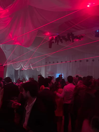 The image depicts a crowded indoor event, likely a party or reception, located in San Luis Potosí City, Mexico. The venue features a high ceiling draped with voluminous white or light-colored fabric, creating a tent-like atmosphere. The entire scene is illuminated predominantly by intense red lighting, augmented by multiple red laser beams crisscrossing the upper space, forming a dynamic grid pattern. Stage lights or spotlights are visible mounted to the ceiling. A visible haze or fog permeates the air, enhancing the laser effects.
Numerous individuals are present, filling the foreground and midground, suggesting a large gathering. Most attendees appear to be adults, generally dressed in semi-formal to formal attire, with men in dark suits or collared shirts and women in dresses. In the immediate foreground, a man in a dark suit jacket and white shirt, seen in profile, holds a clear drinking glass. To his right, another man in similar attire is partially visible from the back. In the midground, a man in a white button-down shirt is observed with his arm around a woman in a light-colored dress (appears orange/pink under red light), viewed from behind. Further into the background, a figure with a distinct horned or pointed headpiece is discernible, silhouetted against a lighter backdrop. Attendees are standing, mingling, and holding beverages.
In the background, two large, white, umbrella-shaped or mushroom-like decorative elements are suspended. The walls of the venue are also draped, featuring circular cutouts or architectural details along their upper section. The overall environment suggests an energetic social gathering.