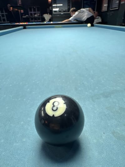 A billiards table, covered in light blue felt, is the primary focus. An eight ball sits prominently on the felt, casting a distinct shadow. A blurred figure of a person holding a pool cue is visible near the edge of the table. The background shows a portion of a bar or entertainment establishment, including tables, chairs, and a wall with framed pictures. There is no information to determine that it is located in Bellville, South Africa.