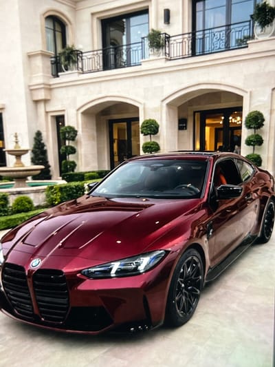 A red BMW M4 is parked on a paved surface in front of a large, modern, light-colored mansion. The mansion has multiple arched doorways, large windows, and balconies with black railings. There is a fountain to the left of the car and manicured bushes and trees surrounding the property. The car is the primary subject, with its hood, grille, headlights, and wheels clearly visible. The sky is not visible in the image.