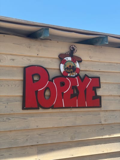 A close-up, angled exterior shot shows a wooden wall with a sign that reads "POPEYE" in large, red, block letters with black and white trim. Above the "POPEYE" sign is a circular decoration resembling a life preserver with a ship's wheel design. Inside the life preserver is a painting of a lighthouse and a silhouette of people. The words "SINCE 1980" and "FILM SET" are also visible on the decoration. The wooden wall is made of horizontal planks, and the background shows a clear blue sky. A blue wooden beam is visible under the roof edge. This sign is located outside the Popeye Village Film Set in Malta.